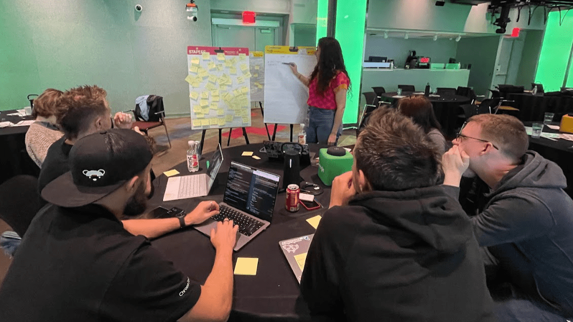 CSS、UI、DevTools のチームが会議室でテーブルを囲んでいます。付箋で覆われたホワイトボードに立つウナ。他のチームメンバーは、飲み物とノートパソコンを置いてテーブルを囲んでいる。