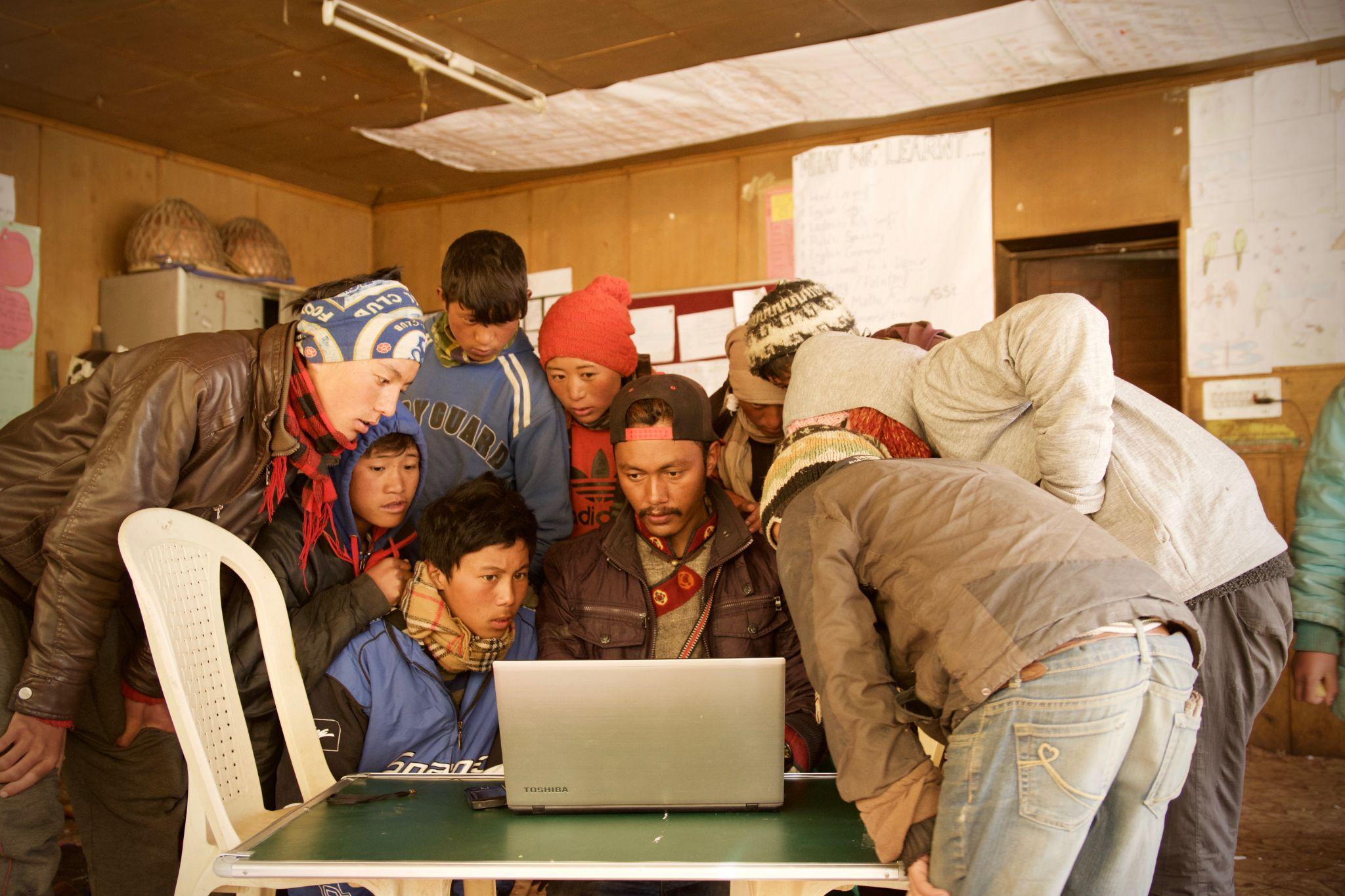 أشخاص يجتمعون حول كمبيوتر محمول يقفون على طاولة بسيطة مع كرسي بلاستيكي على اليسار تبدو الخلفية وكأنها مدرسة في بلد نامٍ.